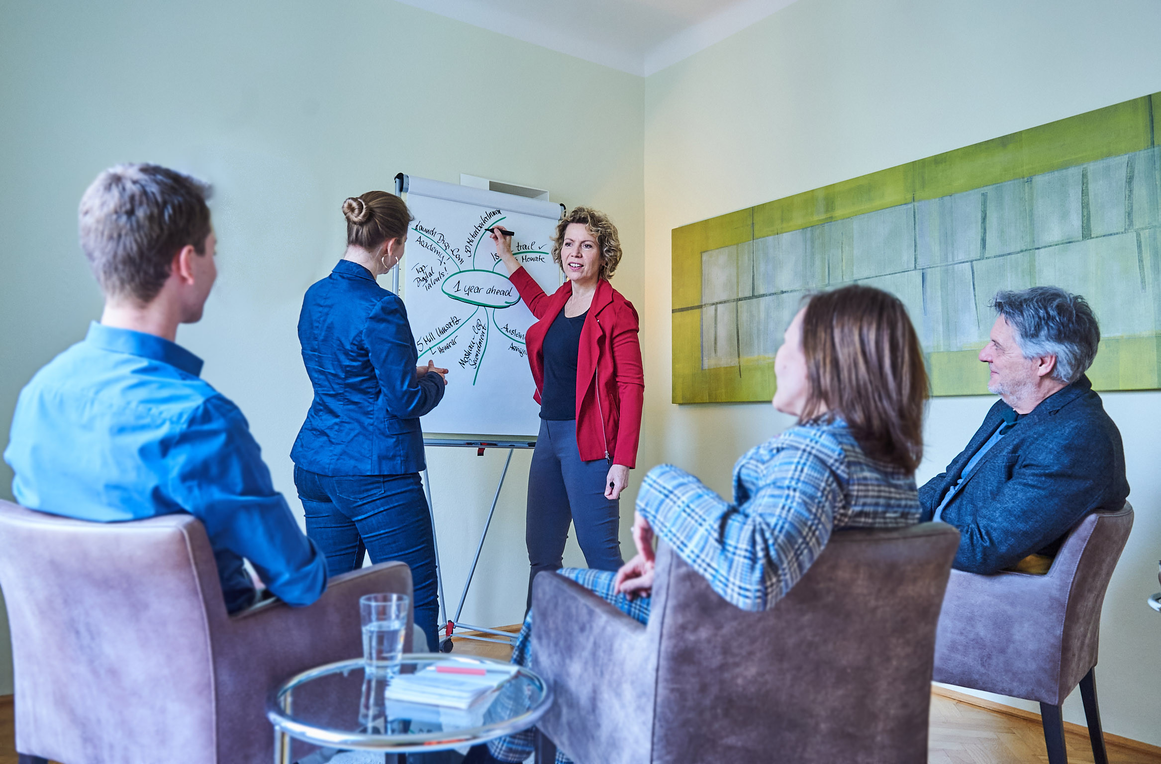 Woman standing at a flip chart with a marker in hand. Explaining her writings to participants sitting around her.