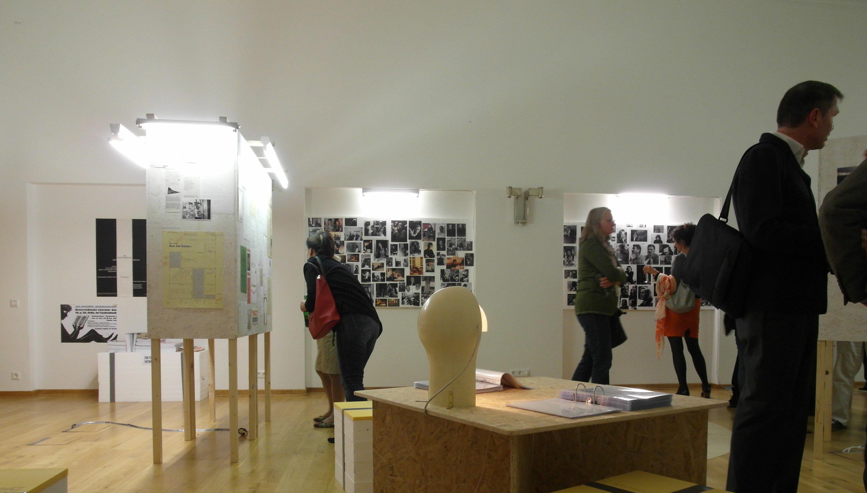 View of Exhibition room: Wooden cuboid element on left side. Sheets of paper with texts and images are mounted on it. Photo pin-wall in the back. Visitors standig around talking and looking at the exhhibits.