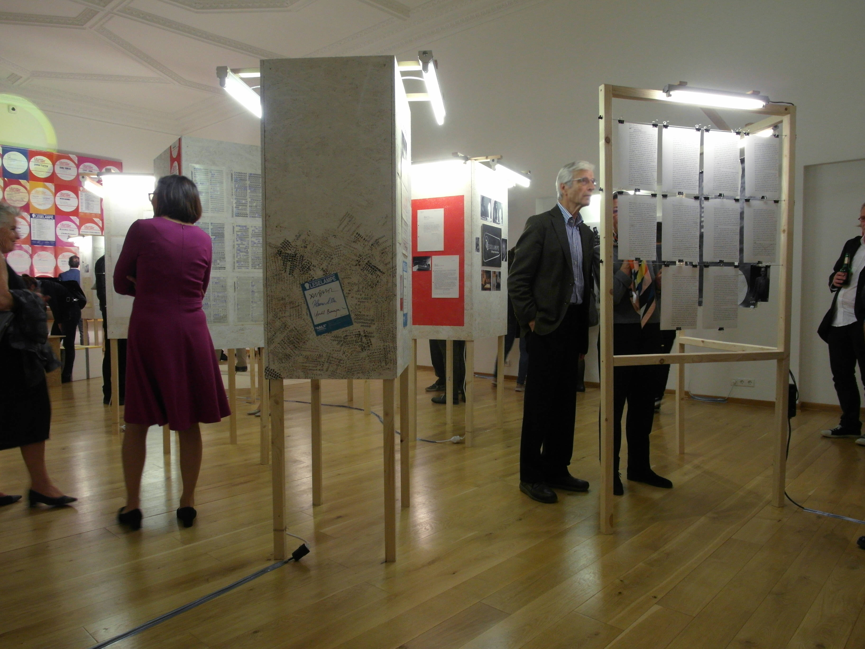 View of Exhibition room: Several wooden cuboids elements. Sheets of paper with texts and images are mounted on the cuboids. Inside a wooden frame metal cords are stretched. Sheets of paper with text are clipped to the cord. Beside visitors talking to each other.