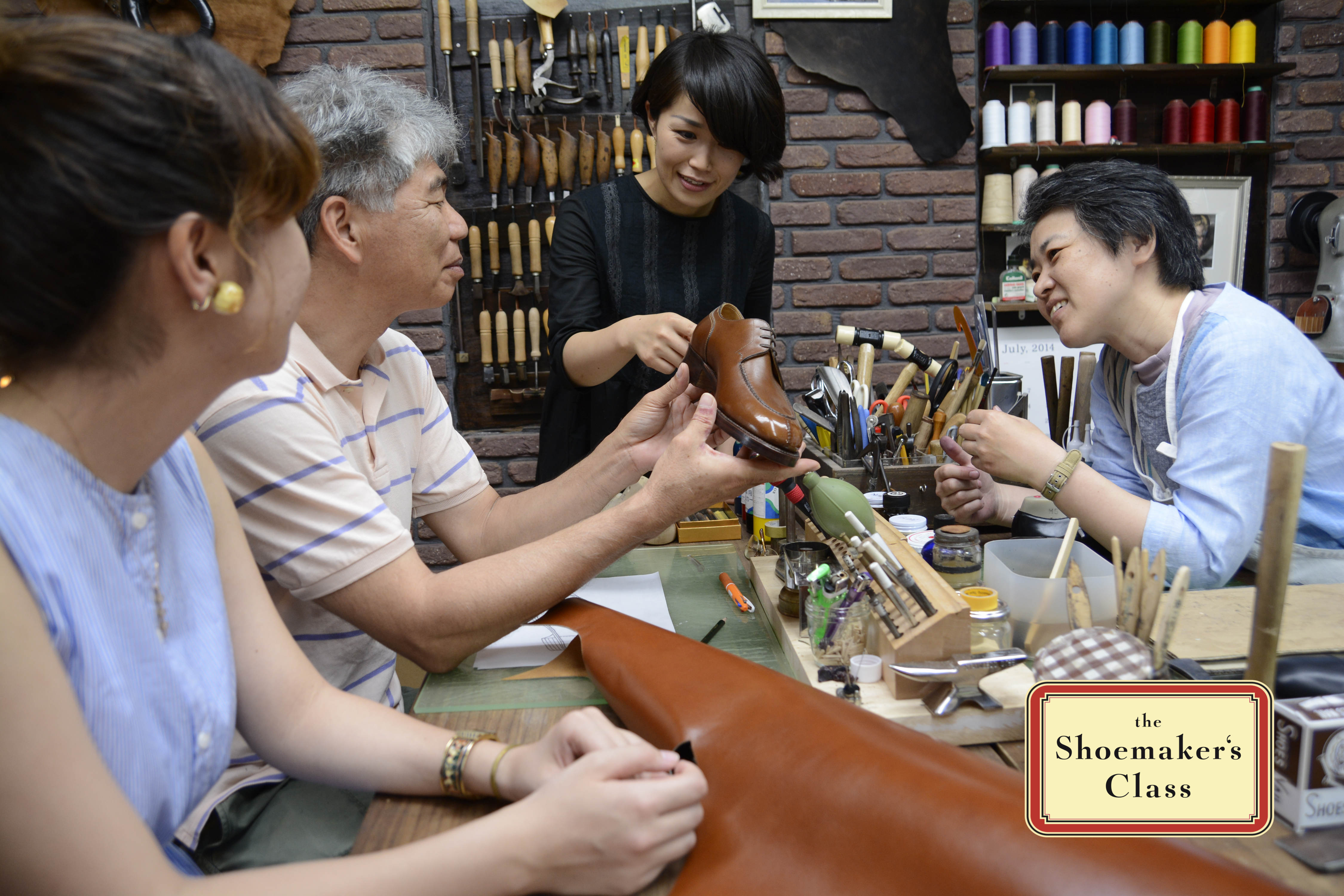 Inside shoemaker's workshop. People sitting around table. One hols shoe. Woman pointing at shoe and explaining.