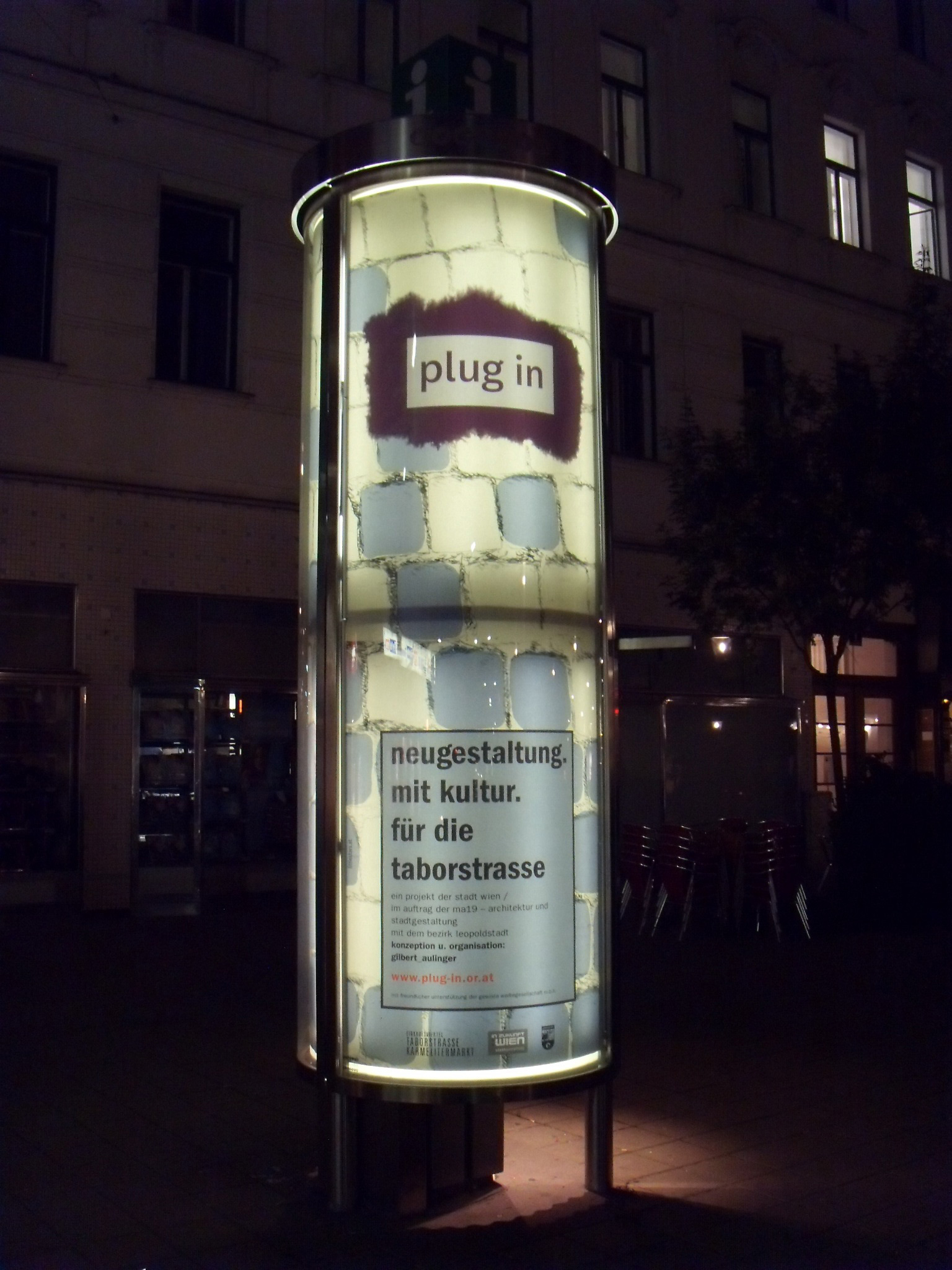 Nighttime. One side of large advertising glass-pillar in the city. Poster illuminated inside.