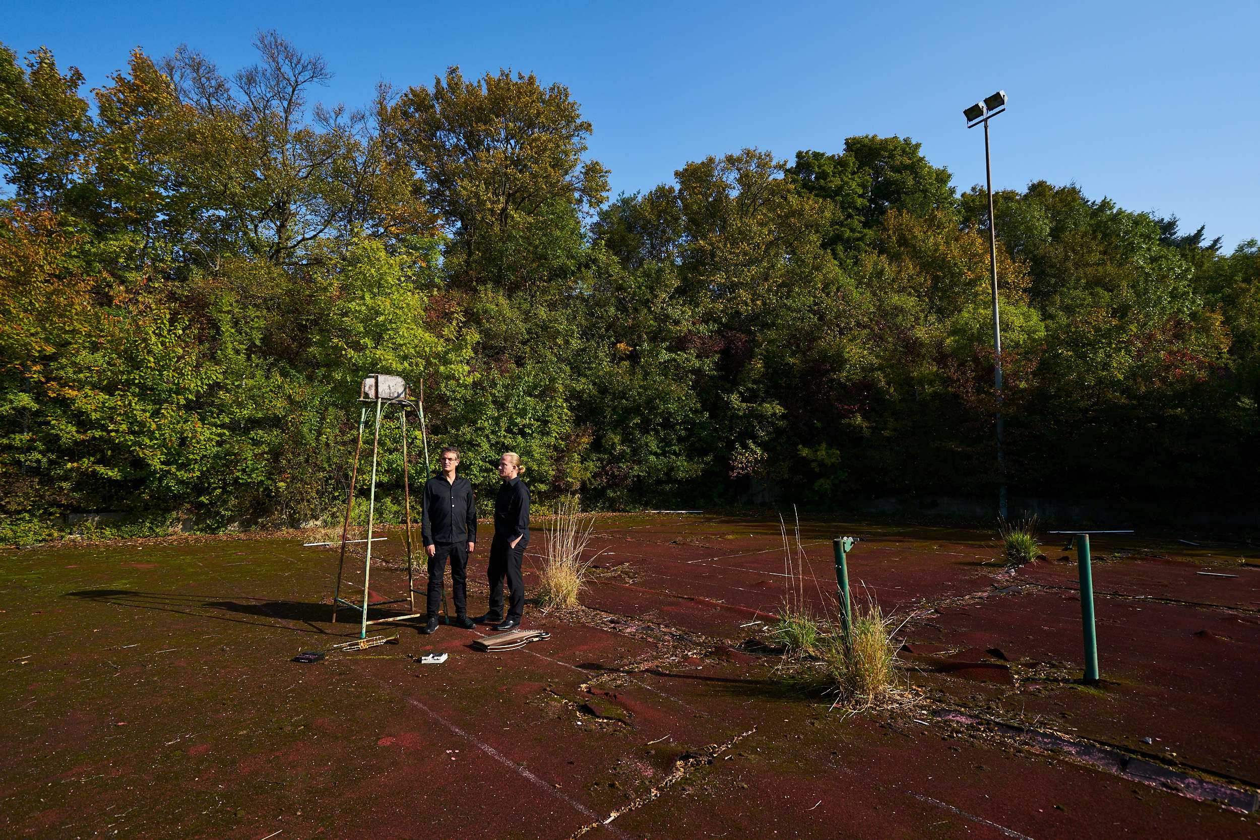 2 man dressed in black standing on an abandoned tennis court.