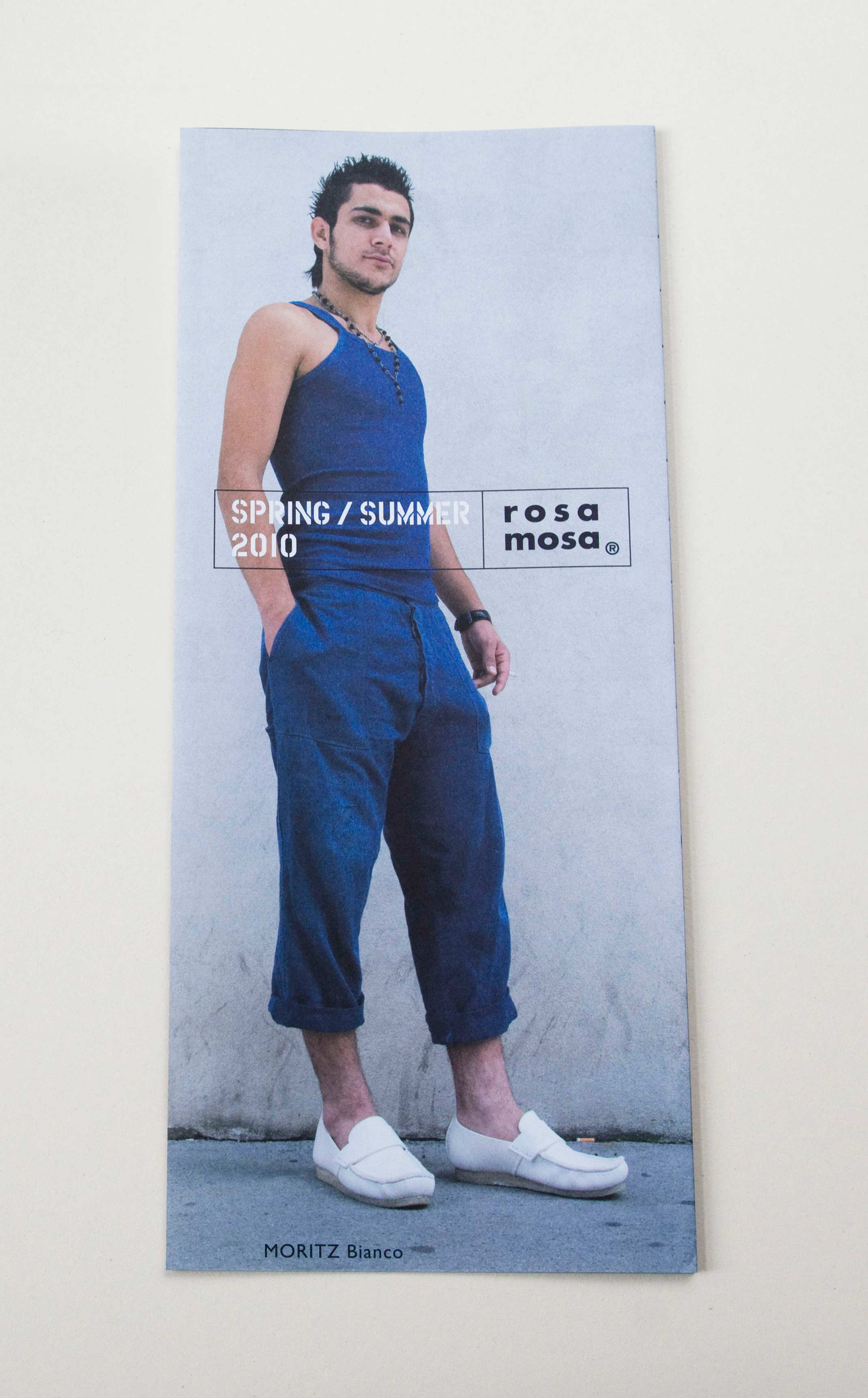 Front of zig-zag folded catalogue. Full-page photo of young man standing on the street in front of white wall. Logo and title in stencil font overlayed in center.