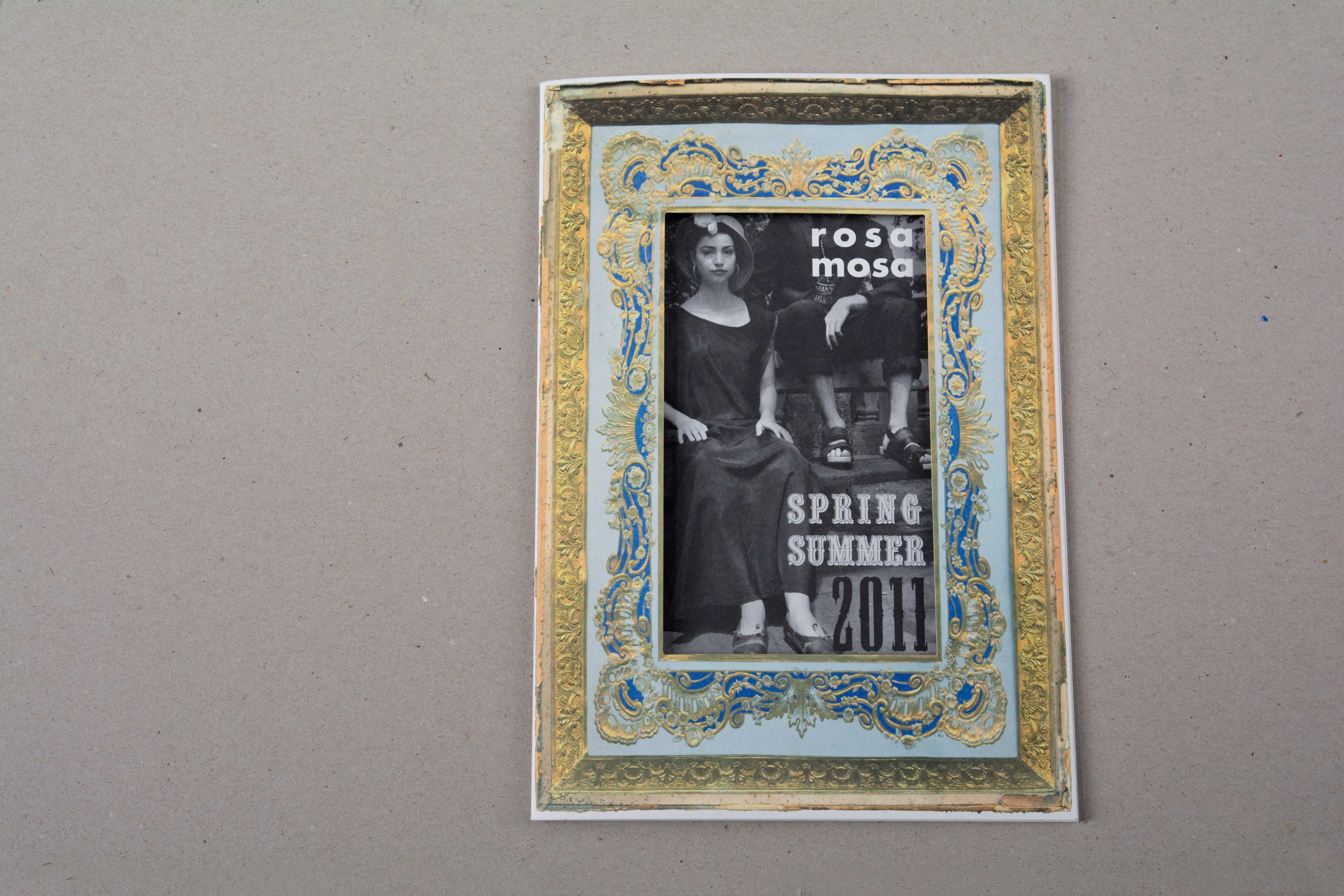 Lookbook cover front. Full-page photo of an old opulent frame with ornaments in gold. In area of inlay a punched rectangle. Partly first page of catalogue is visible. Black/White photo of woman and men sitting on a bench in park. Overlayed by Logo and title in old western style font.