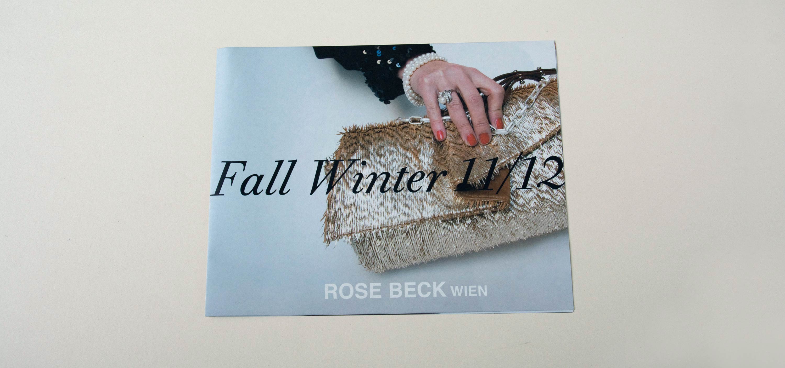 Lookbook cover front. Full-page close-up photo of hand holding handbag. Large title in script font overlayed. Logo at bottom overlayed.