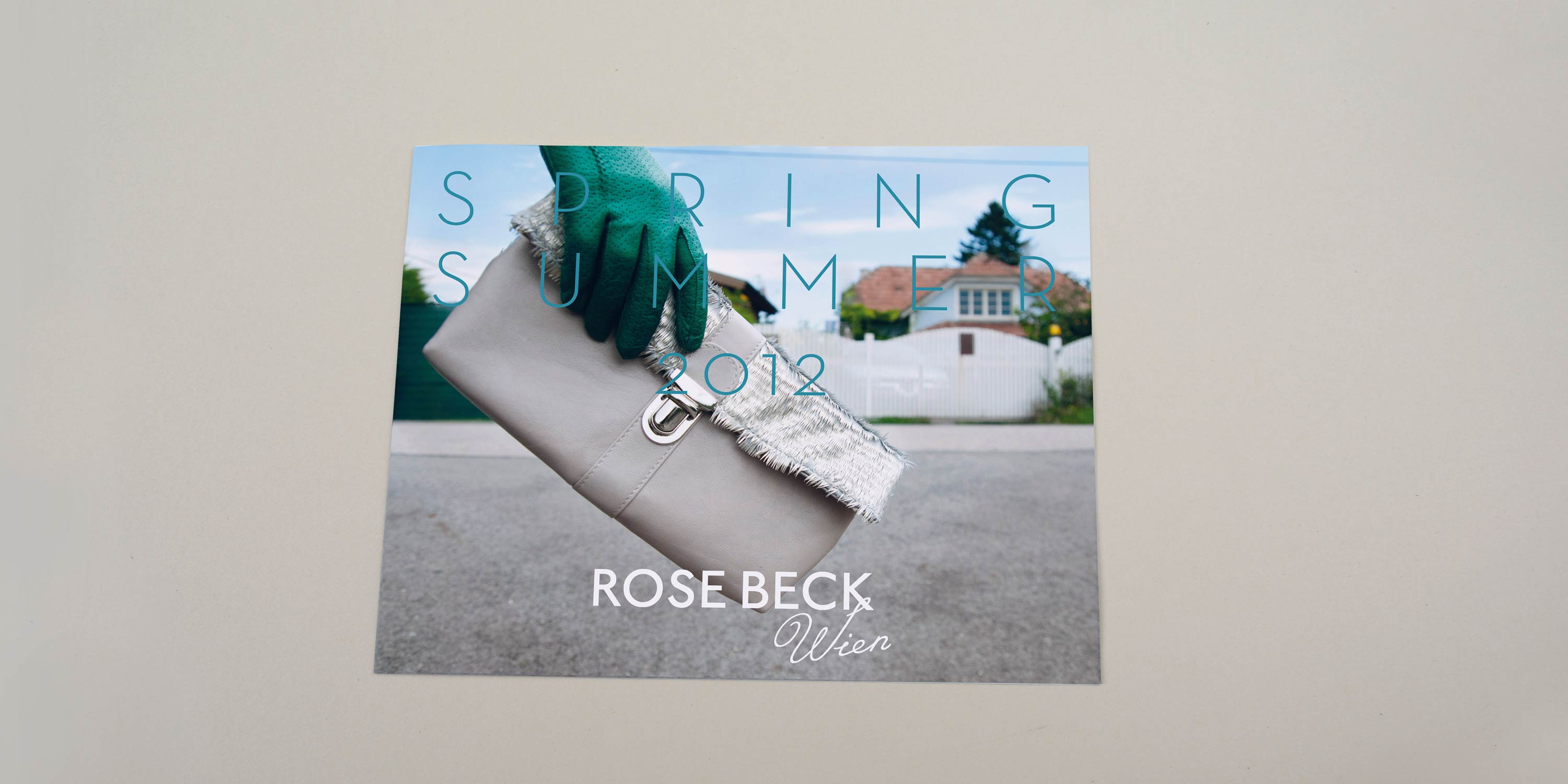 Lookbook cover front. Close-up of hand holding handbag. Fence and houses in the background. Large title font with lots of spacing overlayed. Logo at bottom overlayed.