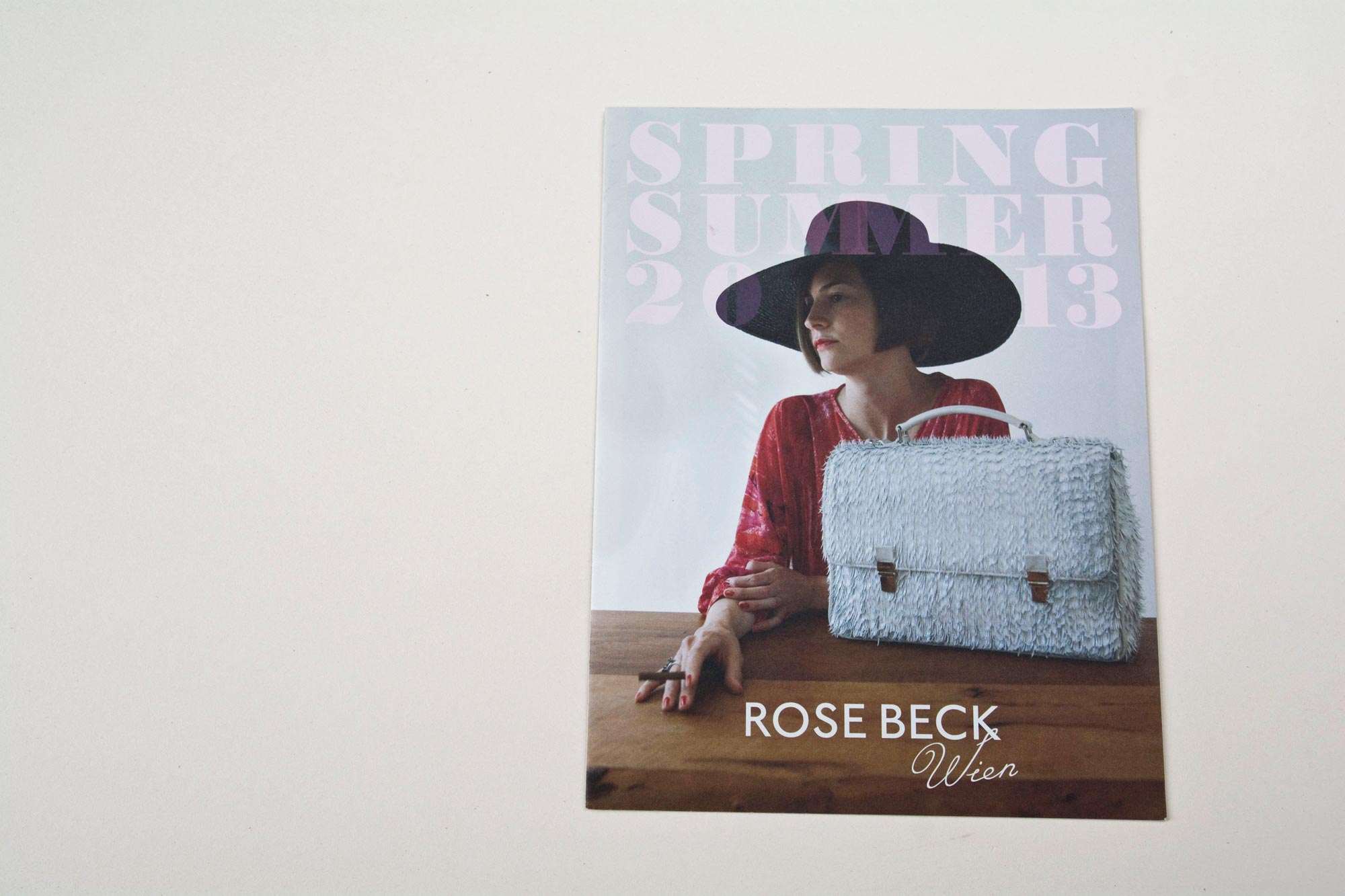 Lookbook cover front. Elegant handbag standing on wooden table. Woman sitting behind. Large title font overlayed. Logo at bottom overlayed.