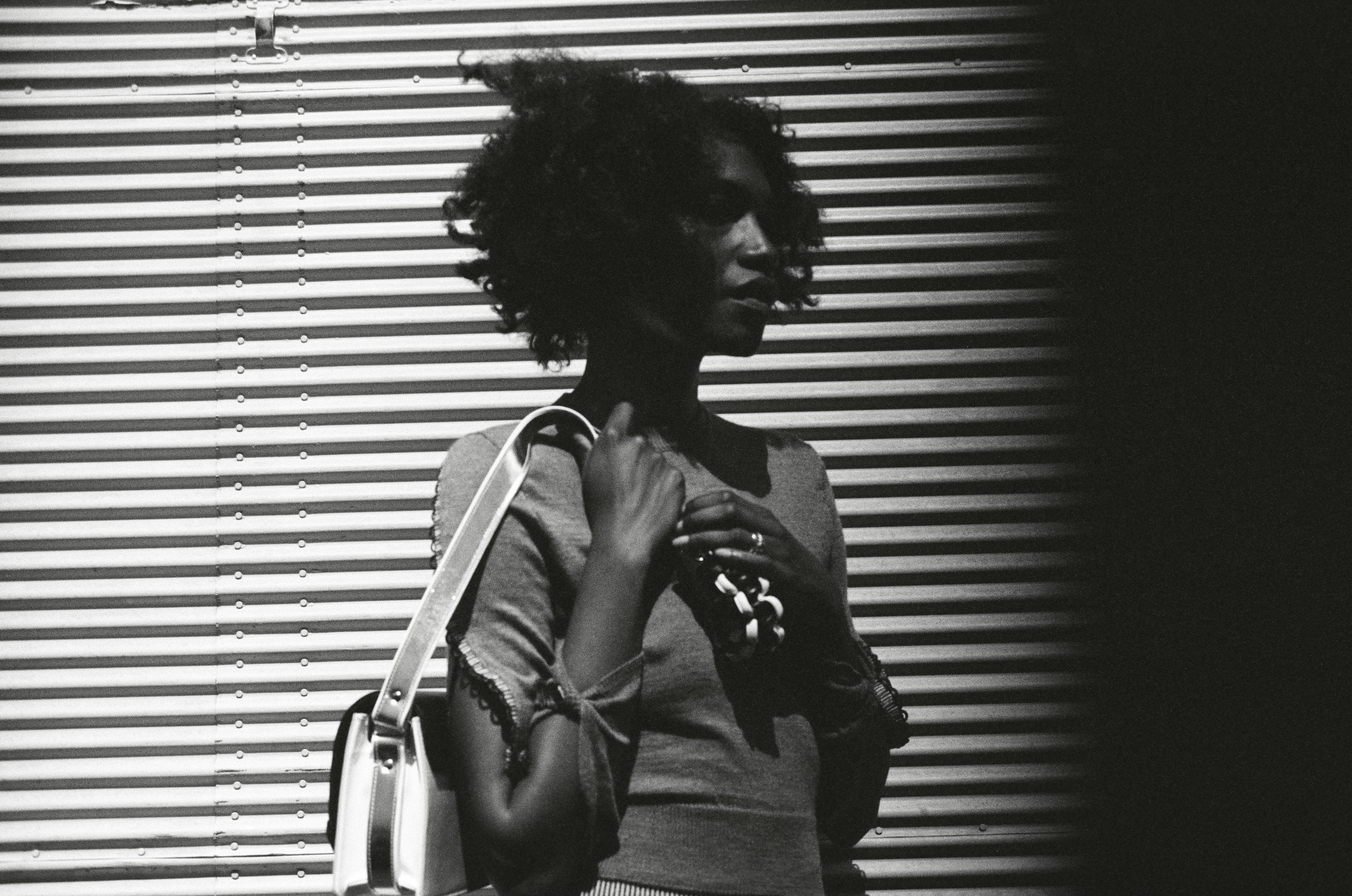 B/W photo. Woman in front of roller shutter. Carries handbag around her shoulder.